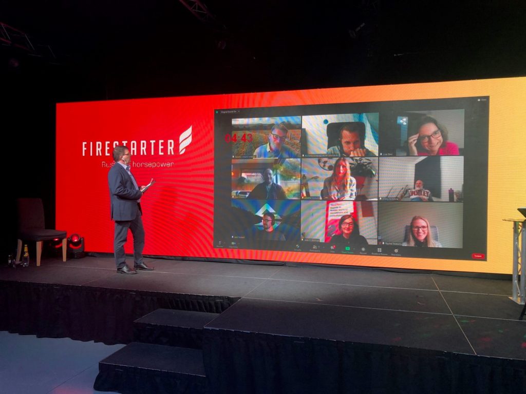 A man an a stage with a large wall screen with a zoom call on it